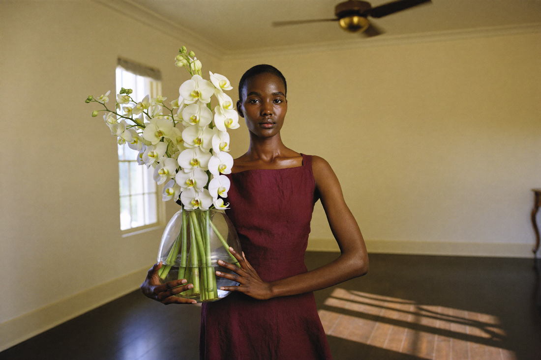 A woman in a dark wine-red dress carries a clear glass vase of tall white orchid sprays across a bare cream room in late afternoon heat, a brass ceiling fan turning above her.