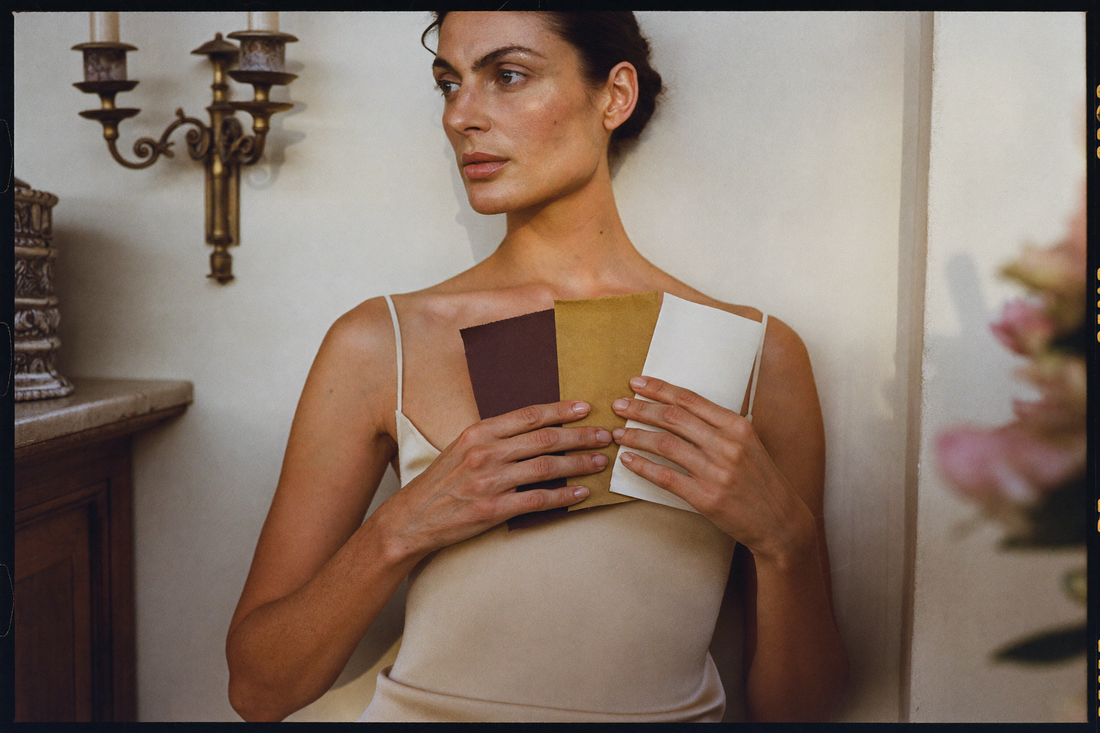 A woman in a cream silk slip holds three sheets of paper against her collarbone, one deep oxblood, one antique gold, one ivory, against a cream plaster wall.