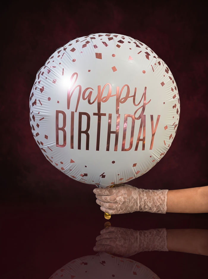 A happy birthday foil balloon in white with rose gold lettering and rose gold confetti, held in a lace-gloved hand