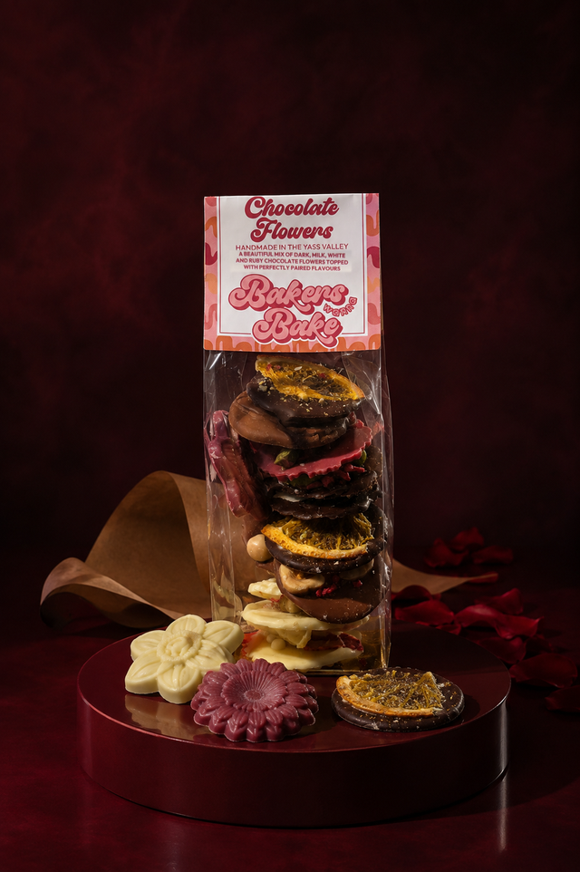Bakers Wanna Bake Chocolate Flowers, a tall clear sleeve of dark, milk, white and ruby chocolate discs, with three loose flowers on an oxblood plinth