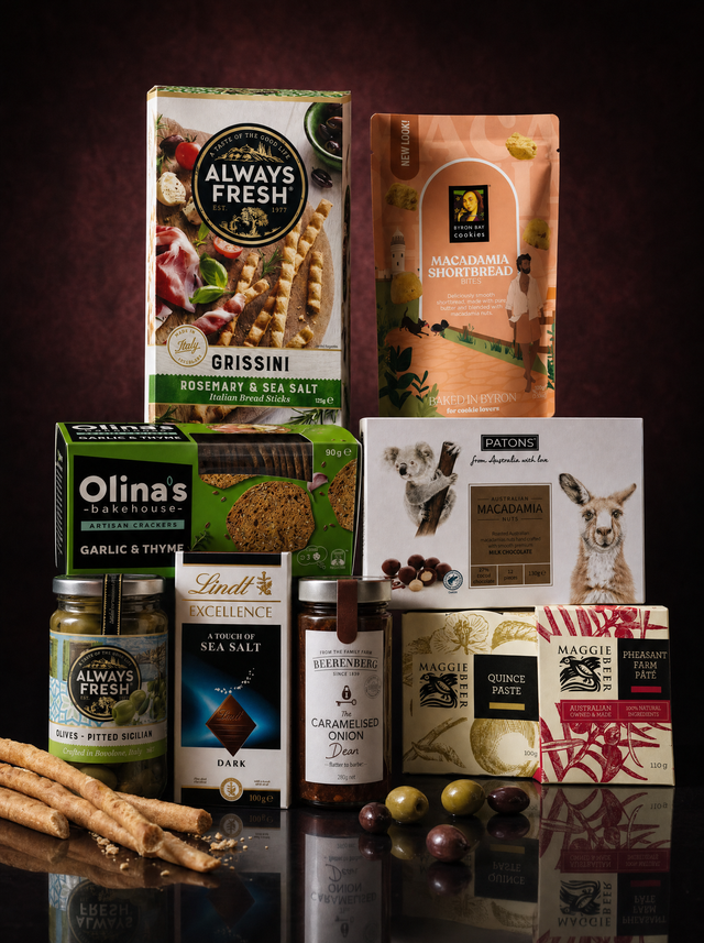 The Classique gourmet hamper from Flowers of Surfers Paradise: Maggie Beer Pheasant Farm Pate and Quince Paste, Olina’s Bakehouse garlic and thyme crackers, Always Fresh pitted Sicilian olives and rosemary grissini, Beerenberg caramelised onion, Patons Australian macadamias in milk chocolate, Lindt Excellence sea salt dark chocolate and Byron Bay Cookies macadamia shortbread, arranged on a dark burgundy background.