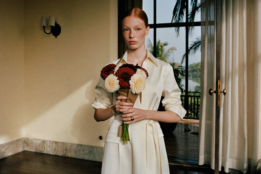 A red-haired woman in an ivory silk shirt dress holds a small hand-tied bouquet of deep burgundy and cream dahlias wrapped in brown kraft paper, in a doorway with palms beyond the window.