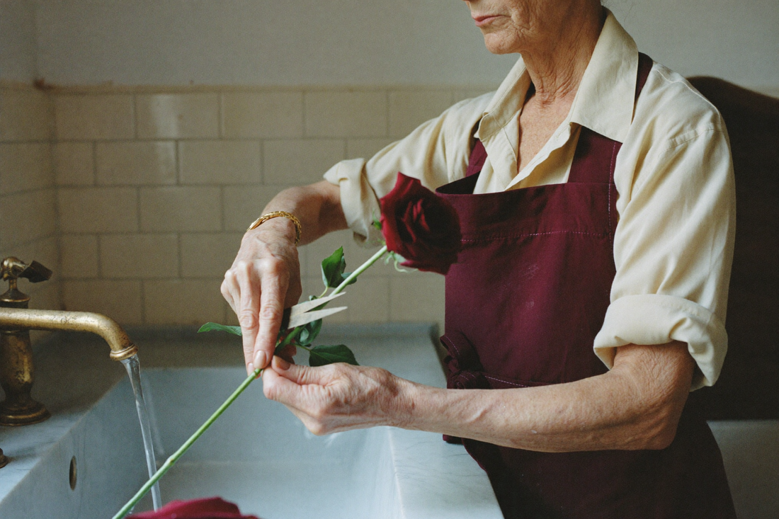 A florist in a cream shirt and wine-red apron trims the stem of a deep burgundy rose at an angle under running water at a white stone sink.