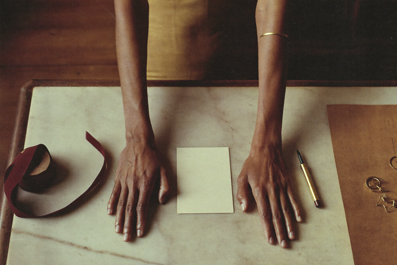 Seen from directly above, a pair of hands rest flat on a marble counter either side of a small blank cream card, with a spool of oxblood ribbon, a gold fountain pen and brown kraft paper beside them.