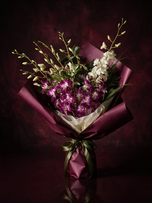 A hand-tied bouquet of magenta and white Singapore orchids with long sprays of green buds, wrapped in deep oxblood paper over an ivory tissue collar and tied with an olive green ribbon