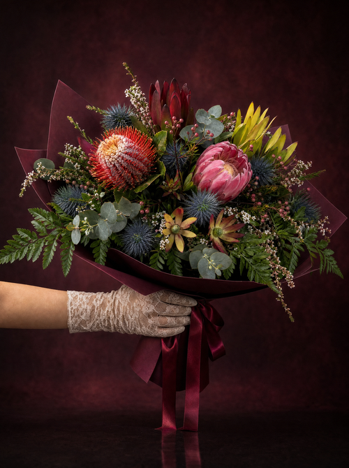 The Native Edit held in a lace glove: pink protea, orange pincushion banksia, blue eryngium and leucadendron in burgundy wrapping, made in Surfers Paradise