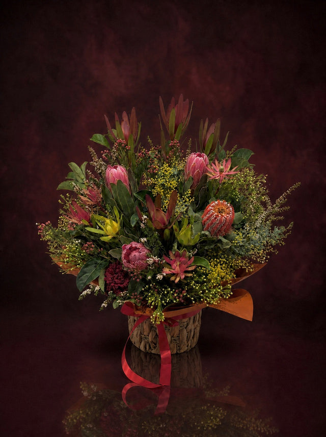 The Native Edit: pink proteas, banksia, leucadendron and wax flower arranged in a woven basket and tied with red ribbon, made in Surfers Paradise
