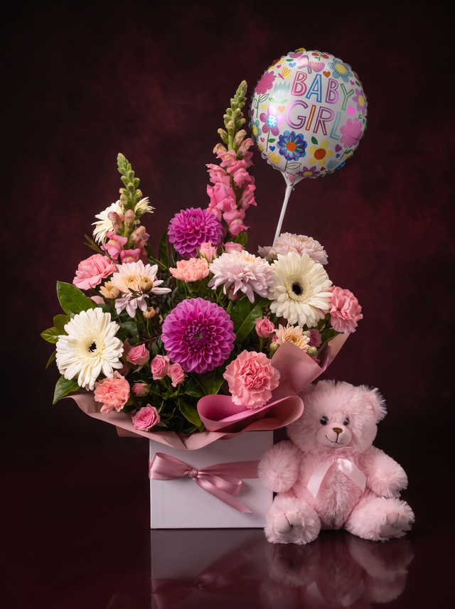 The Nursery in pink: pink dahlias, snapdragons, carnations and cream gerberas in a white box with a pink ribbon, beside a pink teddy bear and a Baby Girl balloon