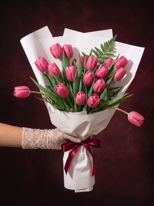 Tulips, the Classique: pink tulips hand-wrapped in white paper and tied with a burgundy ribbon, held in a lace-gloved hand
